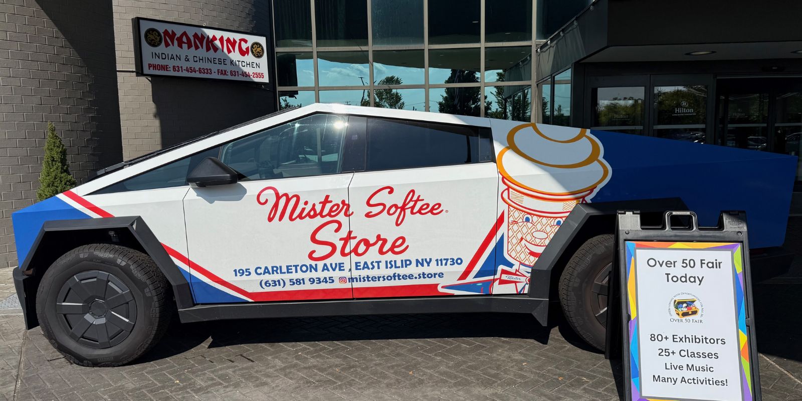 Tesla painted with “Mister Softee Store” is parked in front of a glass building. Nanking Indian & Chinese Kitchen’s sign is also on the building. Next to the Tesla is a sign that announcing “Over 50 Fair Today”