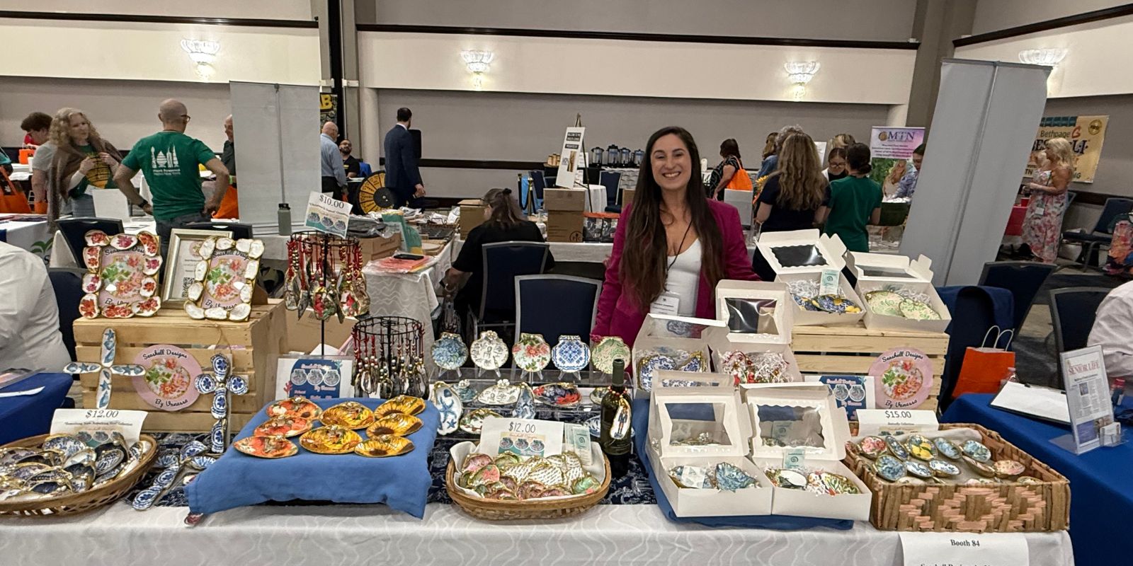 A woman with long brown hair stands behind her table where she is selling colorful painted shells.