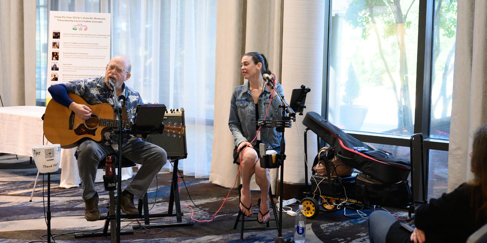 Two singers, one male and one female, sit on chairs while singing a duet. There are some items in the background.