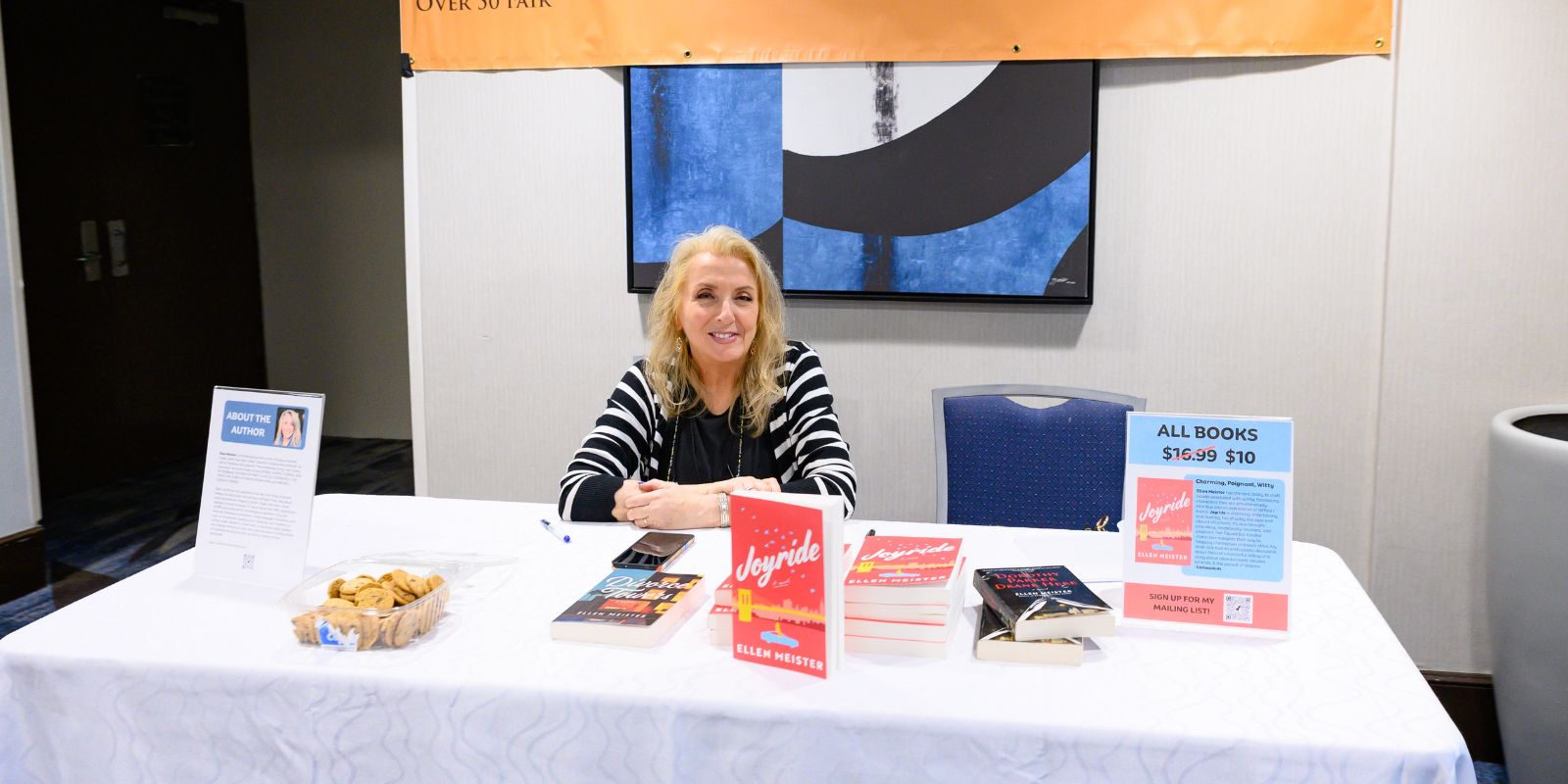 Author Ellen Meister sits at a table selling her books. The titles of the books are Joyride and Divorce Towers. There is a colorful painting behind her.