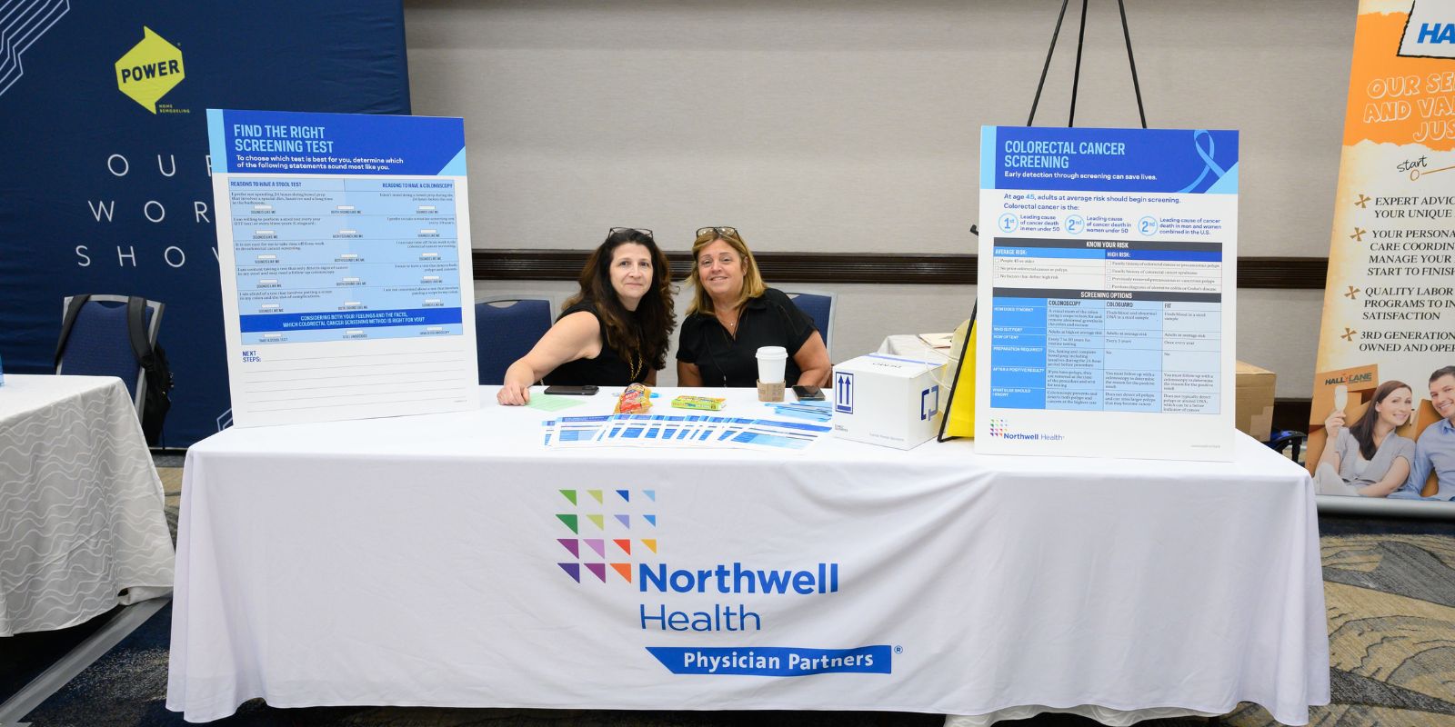 Two women sit at the Northwell Health Physician Partners table with signs about colorectal cancer screenings