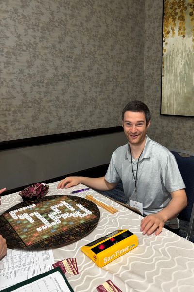 Samuel Kaplan is shown playing Scrabble on a round Scrabble board with ta timer next to him. His opponent is not visible.