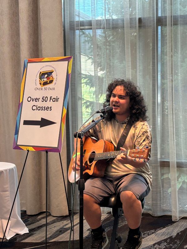 A man in his 20’s sits on a chair while he sings and plays the guitar. Next to him is a sign that reads Over 50 Fair Classes with an arrow